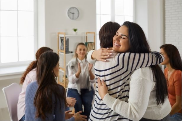 Two women hugging in a room with other women sitting in a circle talking