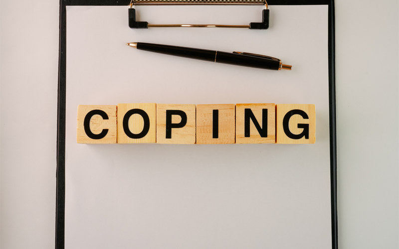 A clip board with a pen and wooden game tiles spelling the word "coping"