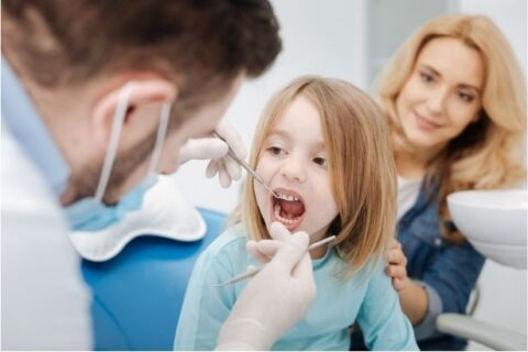 A female child in a dentist chair having their teeth examined by a dentist while the child's mother looks on
