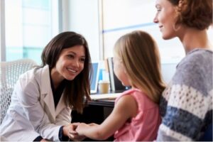 A female pediatrician in a white coat talks to a female child as she sits on her mother's lap