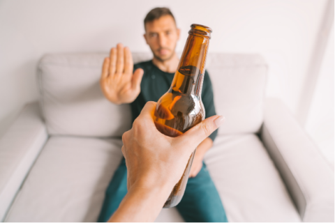 Young white man sitting on a couch holding his hand up saying "no" to a beer being offered