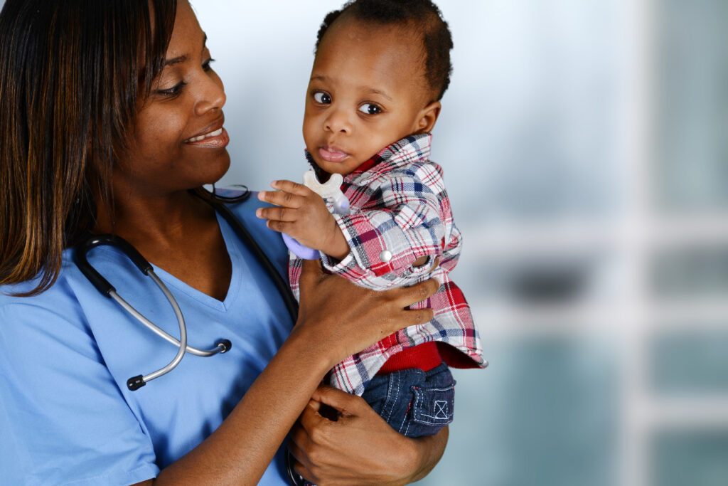 Young African American female healthcare worker in blue scrubs holding a small African American baby. 